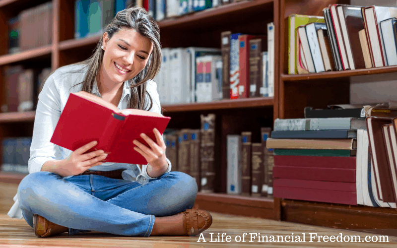 Lady sitting on the floor reading a book with shelves of books behind her. Borrow from the library to help kick start your finances. 