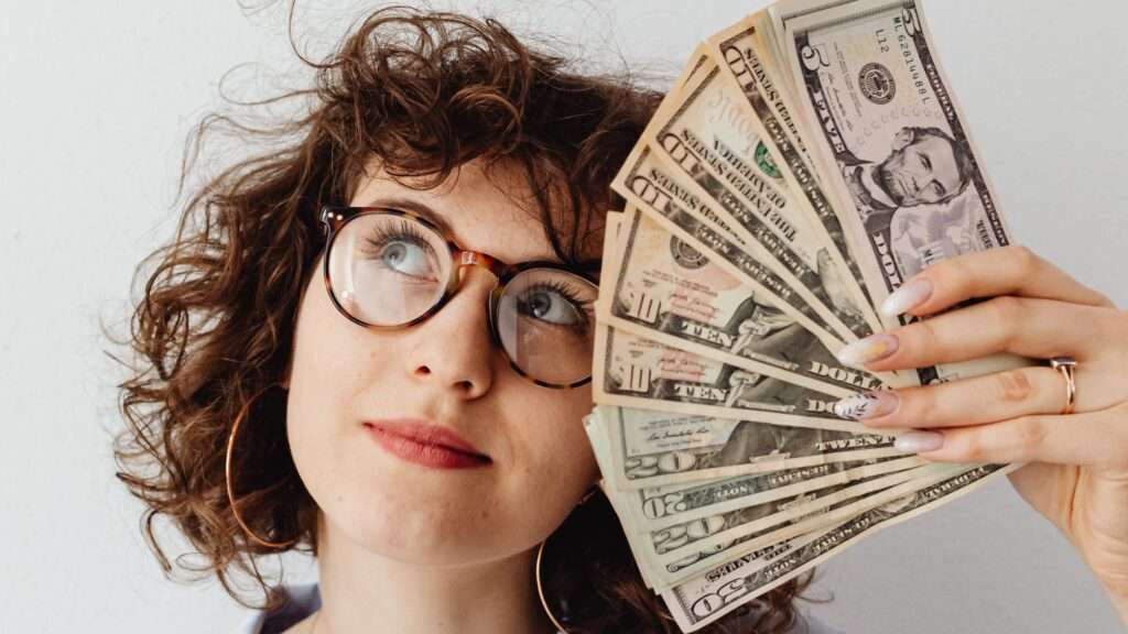 Woman wearing glasses has money spread out in her left hand, holding it up against her face 